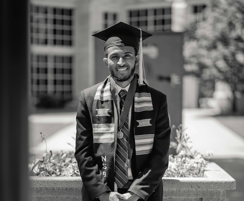 Kujali International Media - Image of a graduate standing in a cap and gown, smiling with his hands folded in front of him.